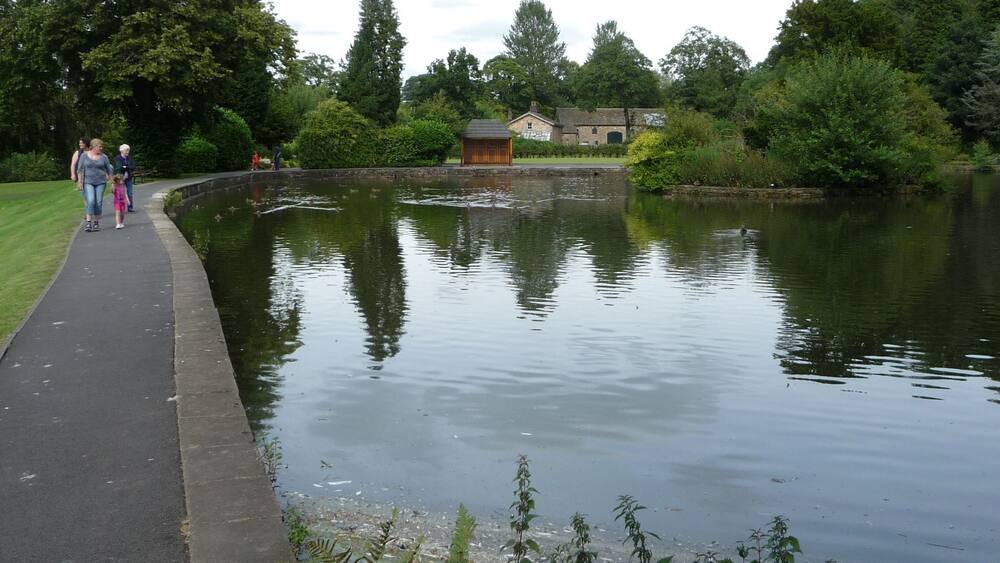 A calm lake with a walking path, surrounded by greenery and a small building.