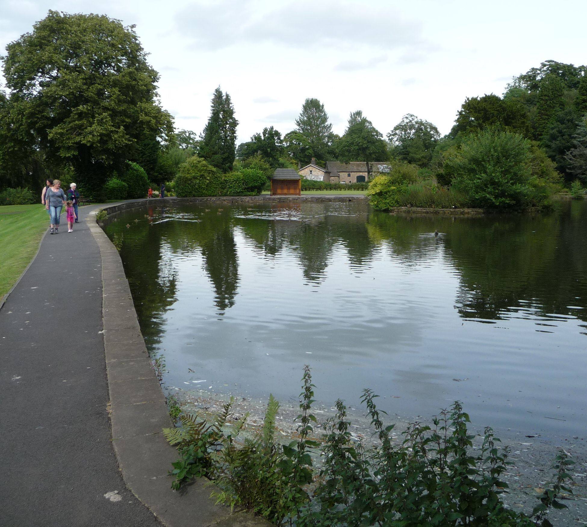 A calm lake with a walking path, surrounded by greenery and a small building.