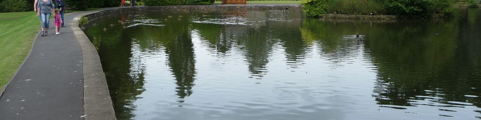 A calm lake with a walking path, surrounded by greenery and a small building.