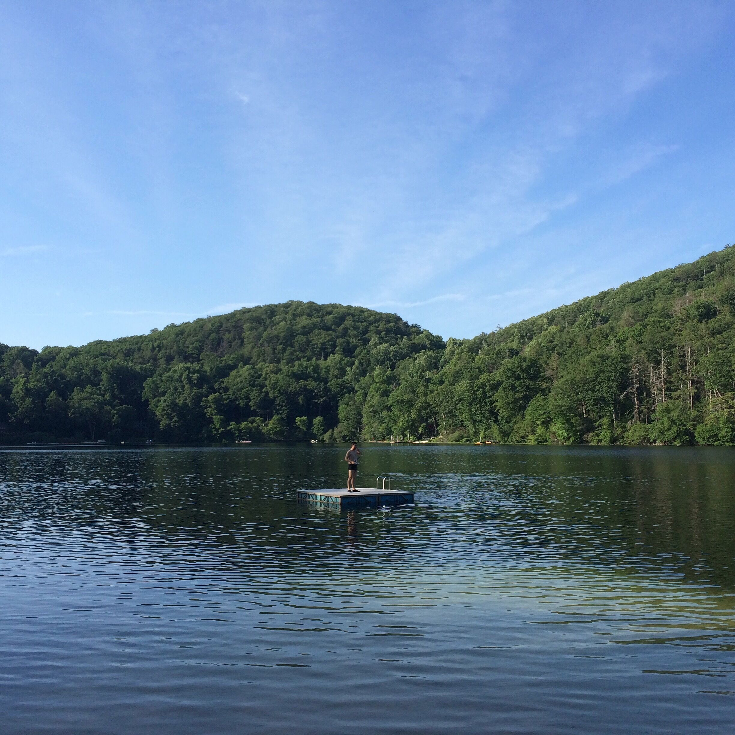 A person standing on a dock extending into a calm lake, surrounded by dense forested hills under a clear sky.