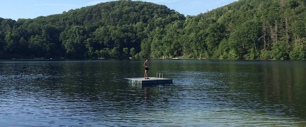 A person standing on a dock extending into a calm lake, surrounded by dense forested hills under a clear sky.