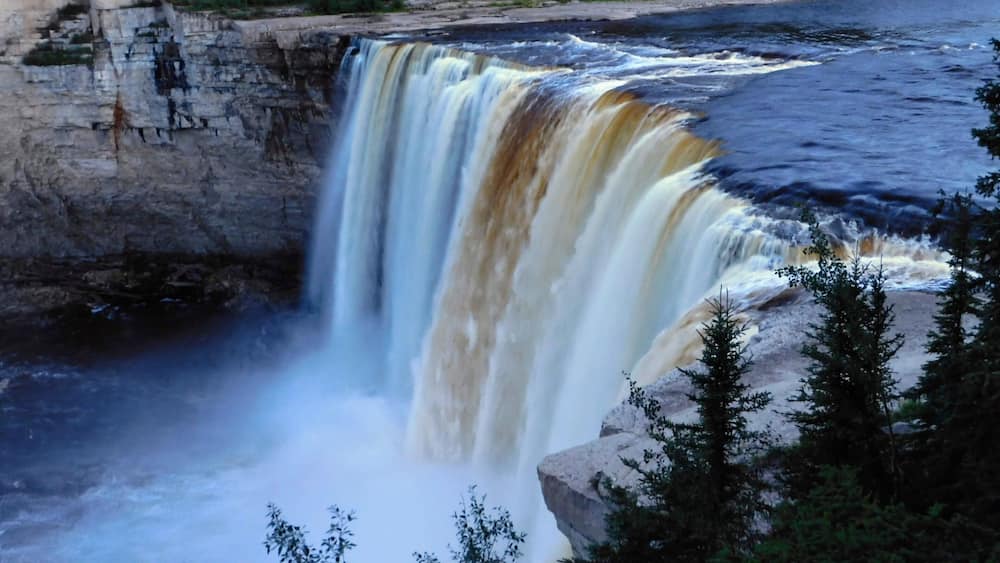 Ein Wasserfall stürzt über eine Felswand in einen Fluss, umgeben von dichtem Wald.