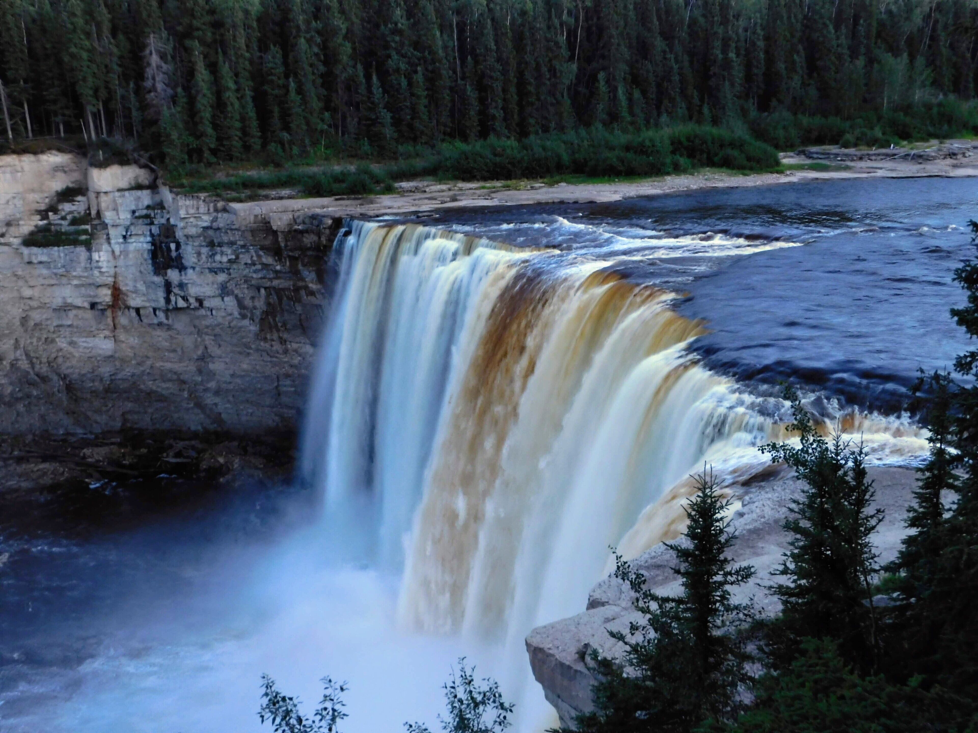 A waterfall cascading over a rocky cliff into a river, surrounded by dense forest.