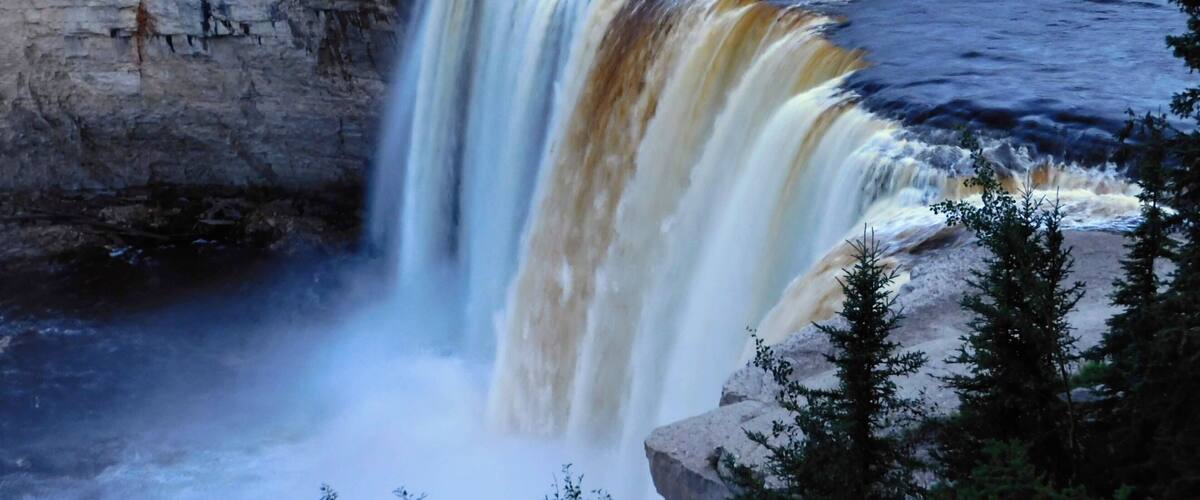 A waterfall cascading over a rocky cliff into a river, surrounded by dense forest.