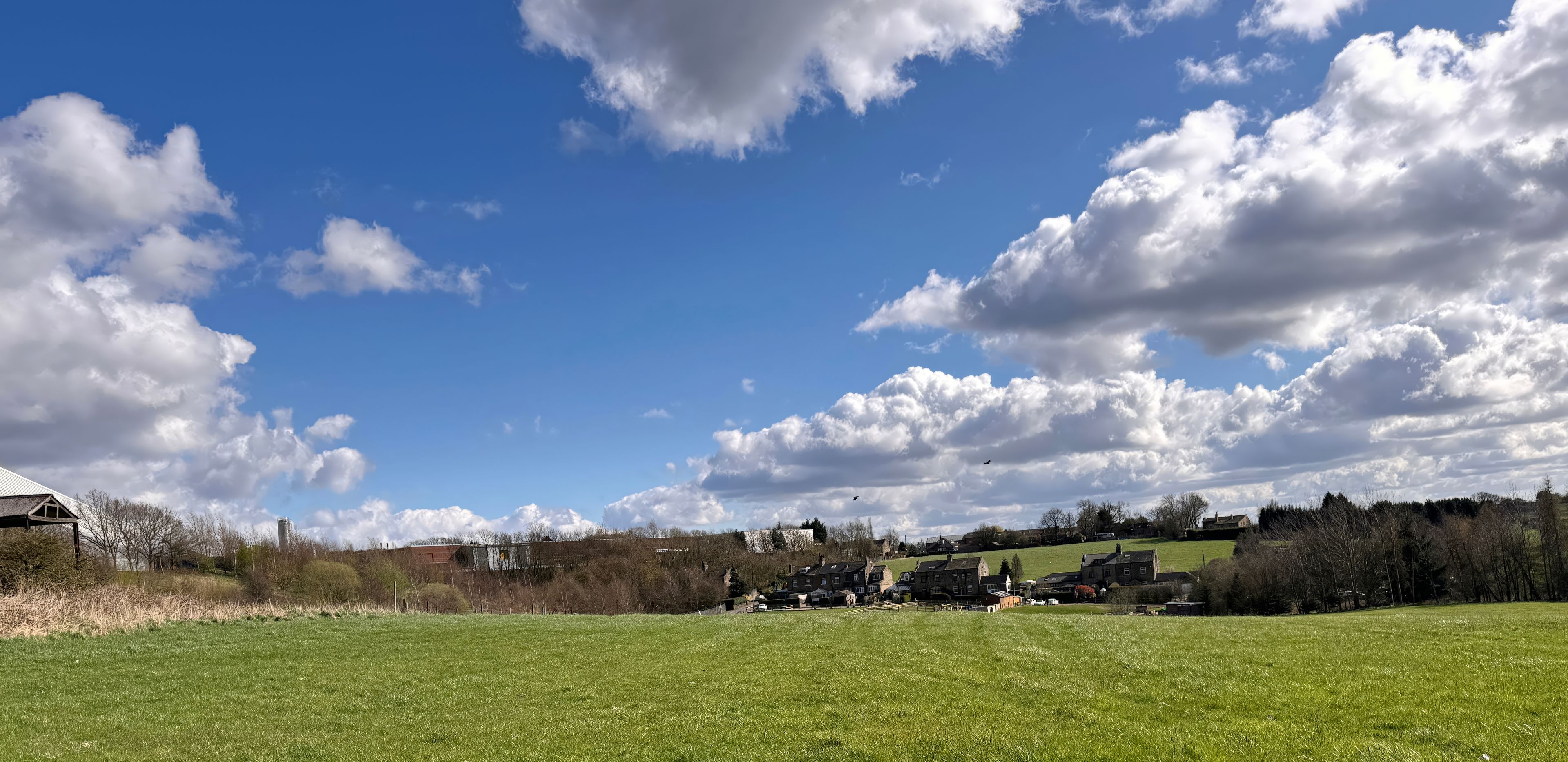 A rural landscape with a clear blue sky, scattered clouds, and a green field.
