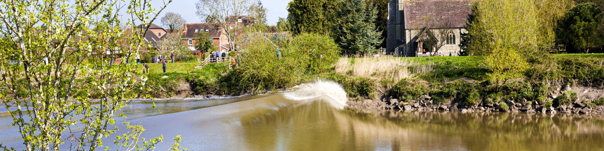 Un río con una pequeña cascada, rodeado de vegetación y una iglesia al fondo.