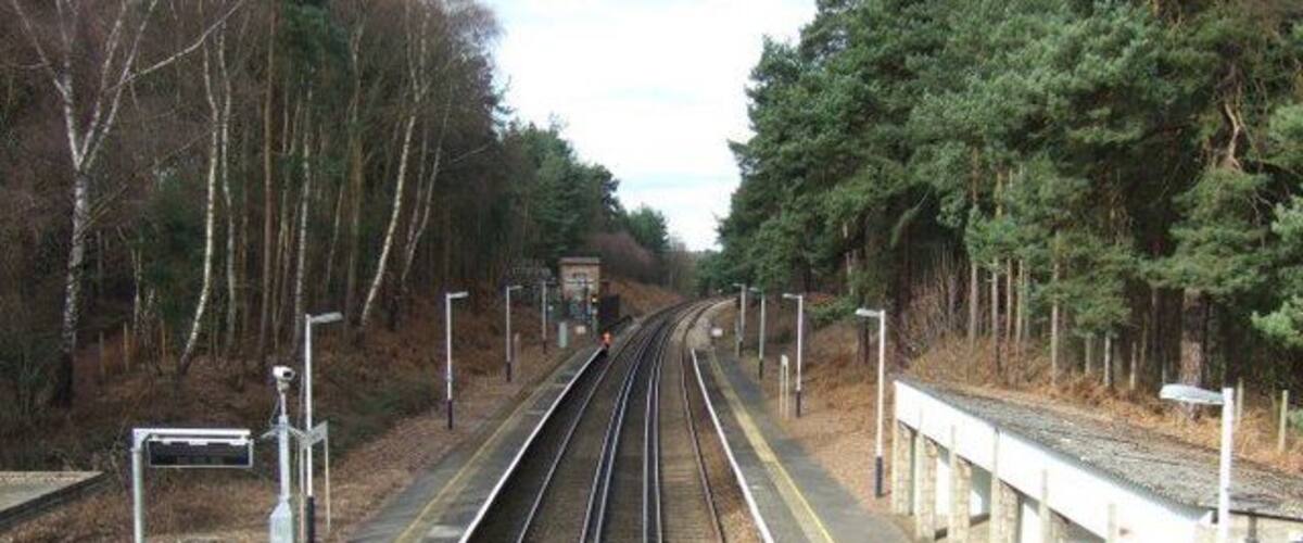 A railway station platform with tracks, a fence, and trees in the background.