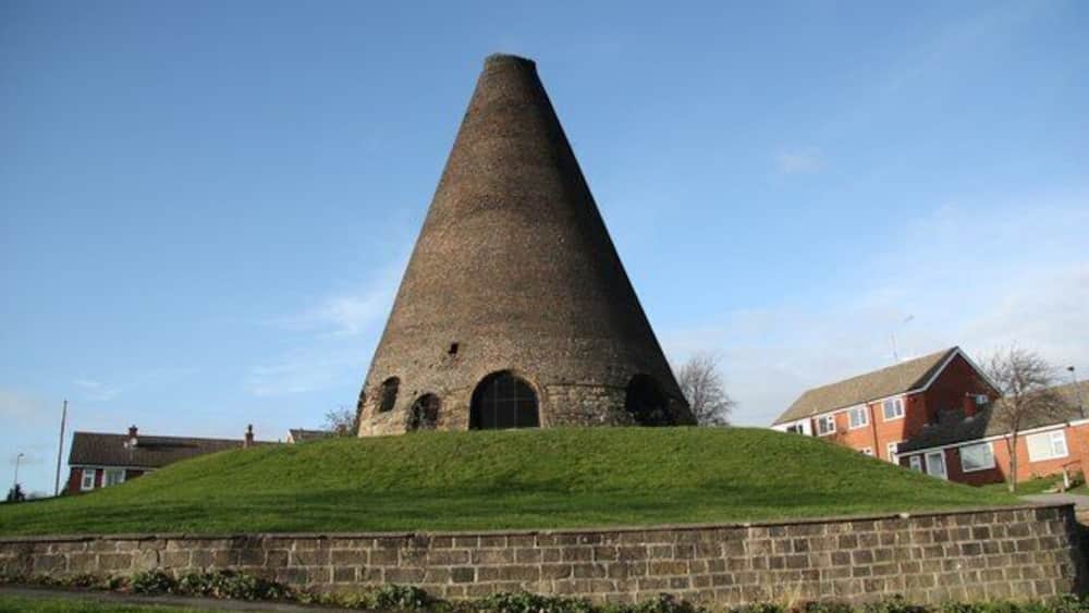 Une structure conique en pierre, située sur une colline herbeuse, avec un mur de briques au premier plan et des maisons en arrière-plan.
