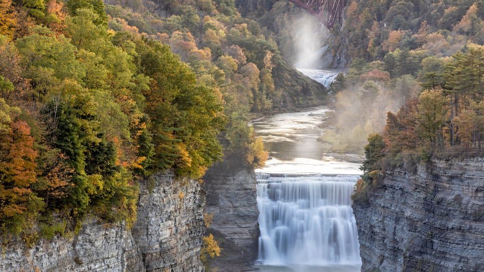 A waterfall cascading down a rocky gorge, surrounded by autumn foliage and a bridge in the background.