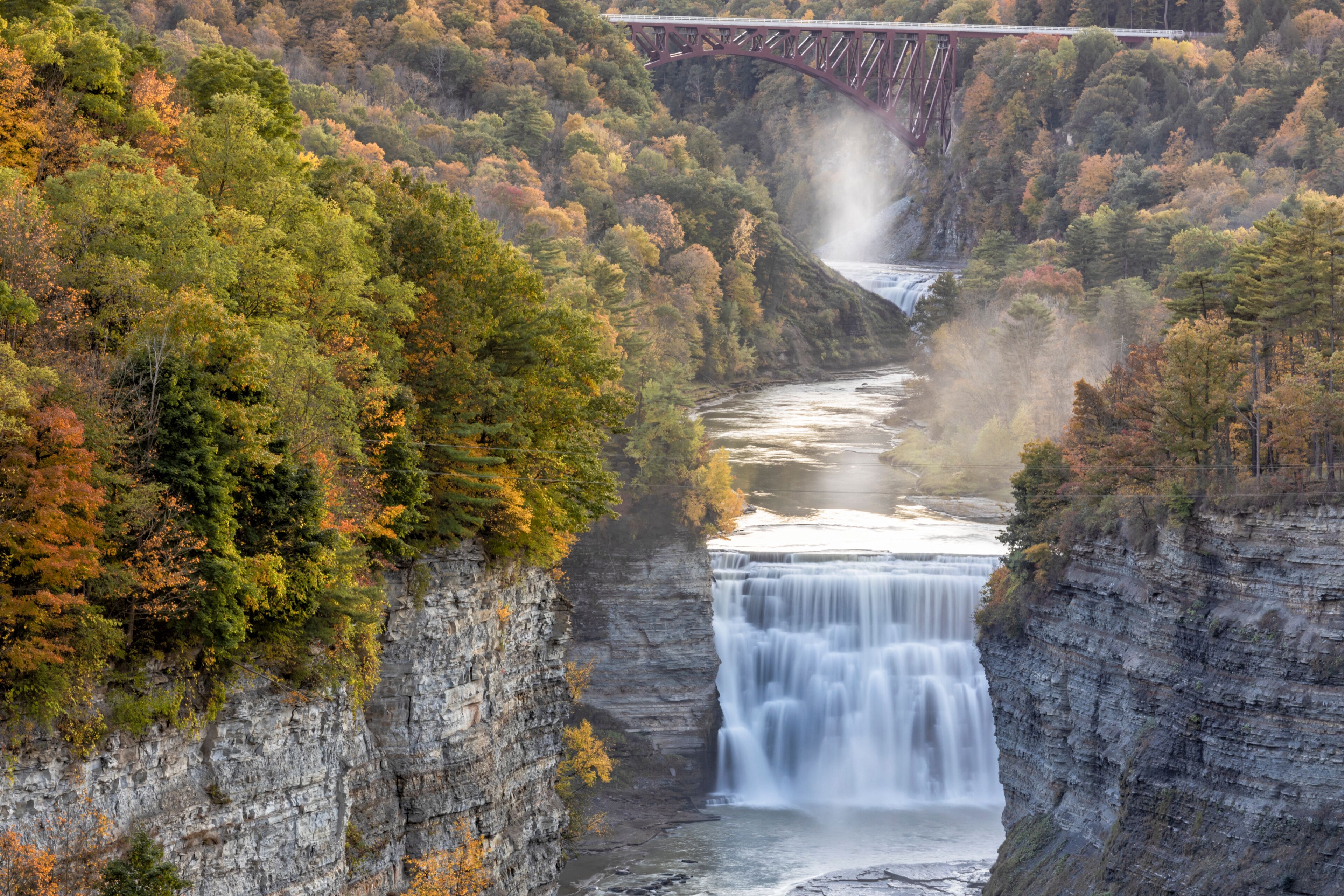 A waterfall cascading down a rocky gorge, surrounded by autumn foliage and a bridge in the background.