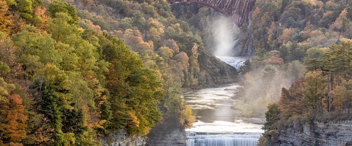 A waterfall cascading down a rocky gorge, surrounded by autumn foliage and a bridge in the background.