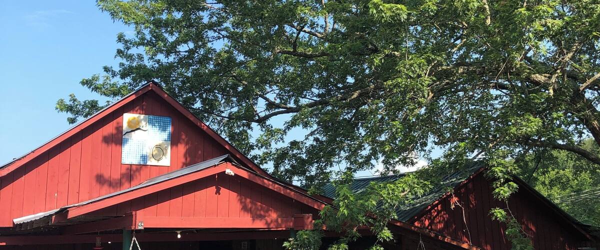 A red wooden building with a covered porch, surrounded by greenery and a large tree.