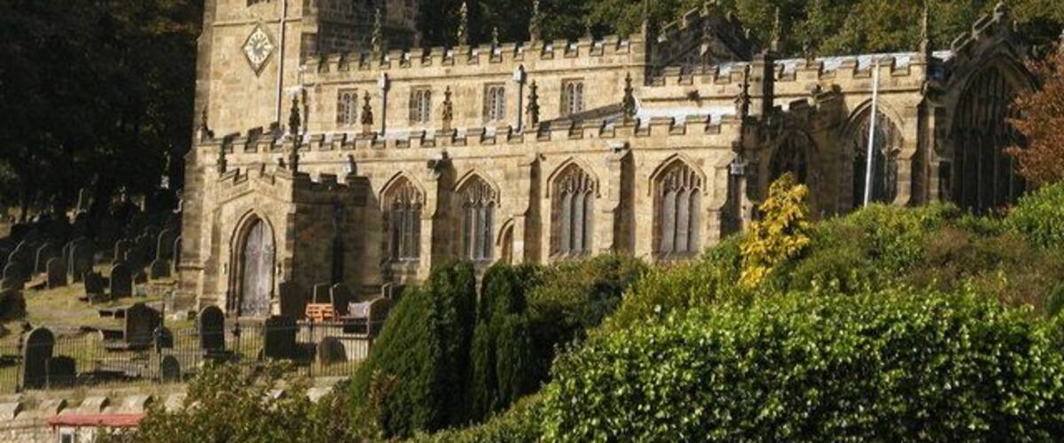 Une église historique en pierre, dotée d’un clocher imposant et de fenêtres en arc de cercle, entourée d’un cimetière et d’une végétation luxuriante.