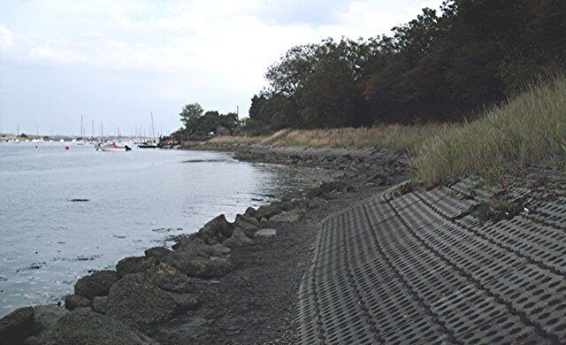 A rocky shoreline with a patterned walkway leading to the water, boats in the distance, and dense trees along the shore.