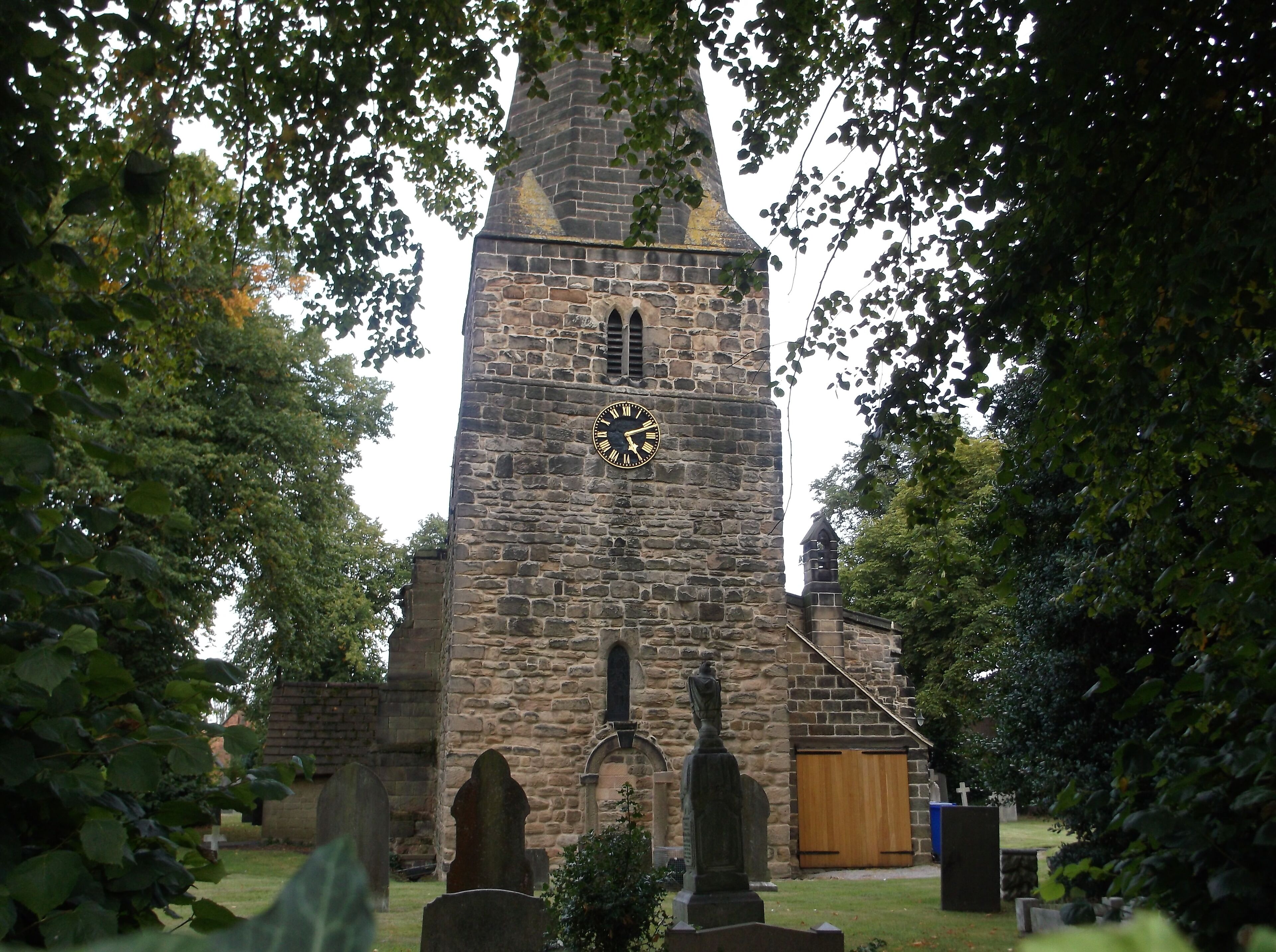 A stone church with a clock tower, surrounded by trees and a cemetery.