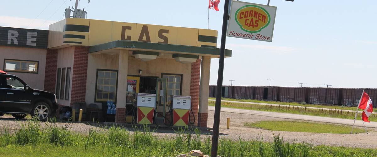 A gas station named 'CORNER CAS' with a Canadian flag, a black truck parked in front, and a fence in the background.