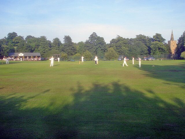 A cricket field with players, a church, and trees in the background.