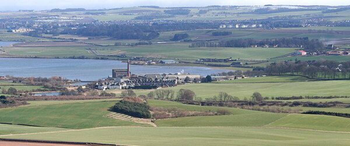 A rural landscape with a large body of water, green fields, and a distant industrial area with smokestacks.
