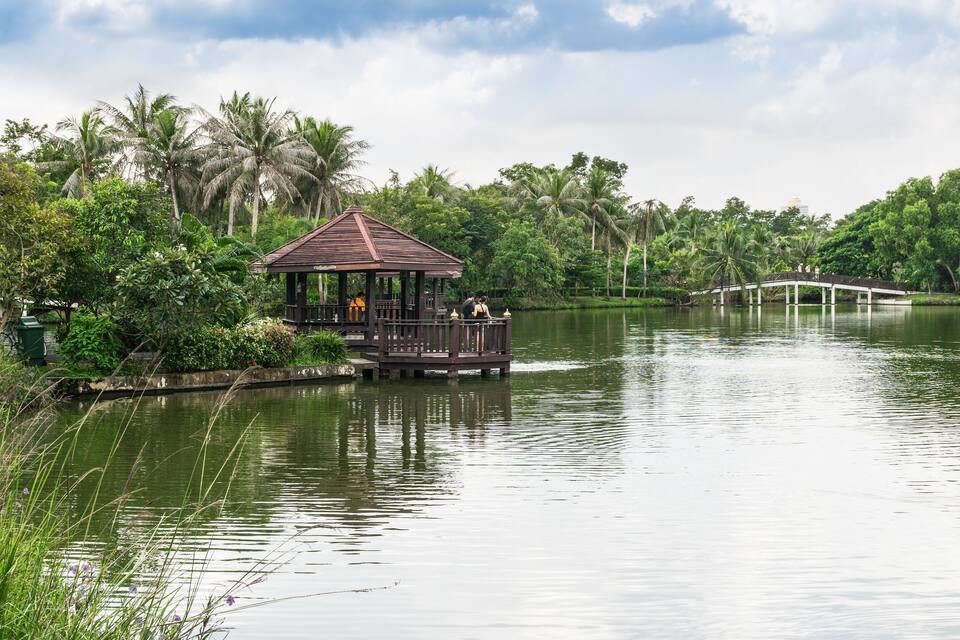 A wooden gazebo on a lake surrounded by lush greenery and palm trees.
