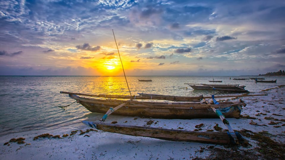 A sunset over a beach with several wooden boats anchored in the sand.
