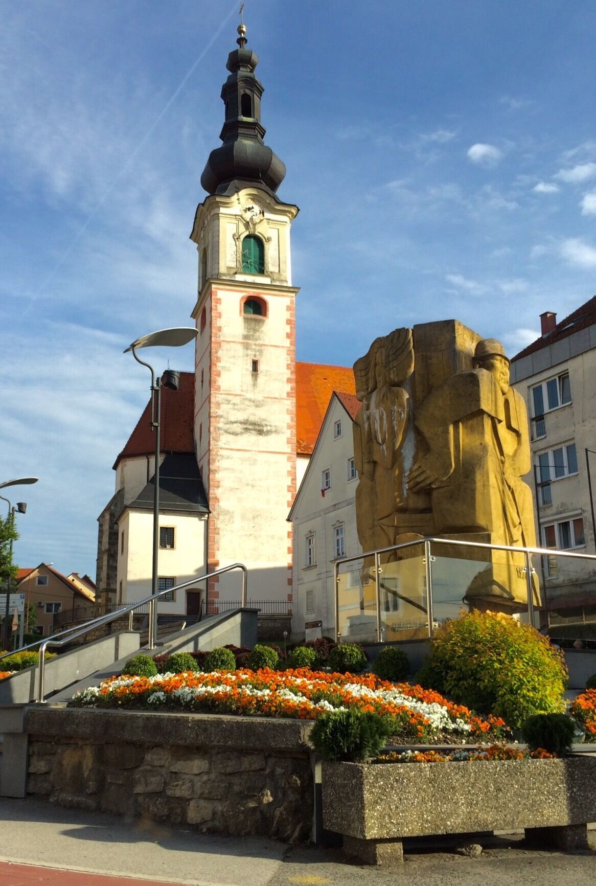 A church tower with a clock, a statue, and flower beds.