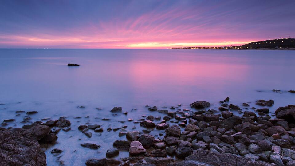 A rocky shoreline at sunset with a calm sea and a city skyline in the distance.