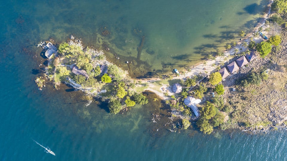 An aerial view of a coastal area with clear water, a boat, and a series of beach umbrellas and huts along the shoreline.