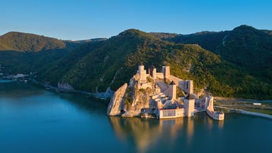 Um castelo medieval numa ilha rochosa, rodeado por um lago calmo e colinas arborizadas.