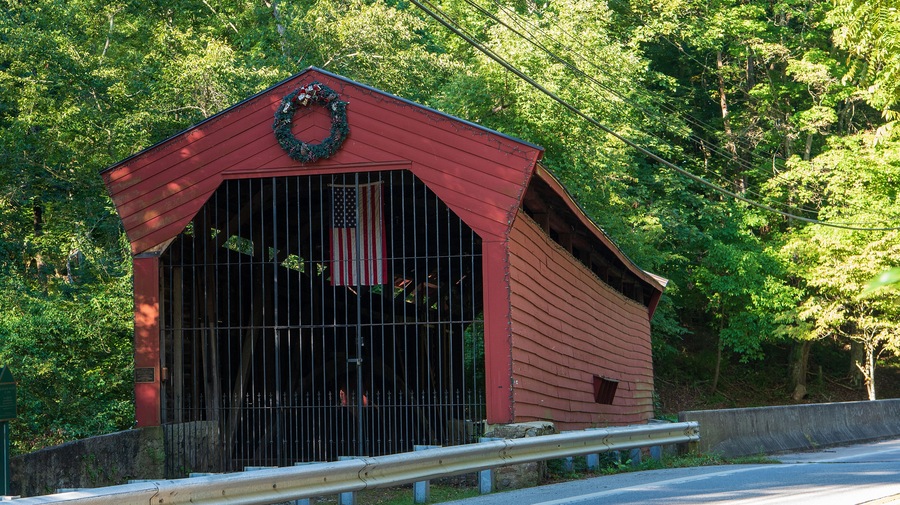 A red covered bridge with a wreath on top, surrounded by lush green trees.