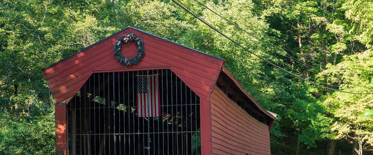 A red covered bridge with a wreath on top, surrounded by lush green trees.