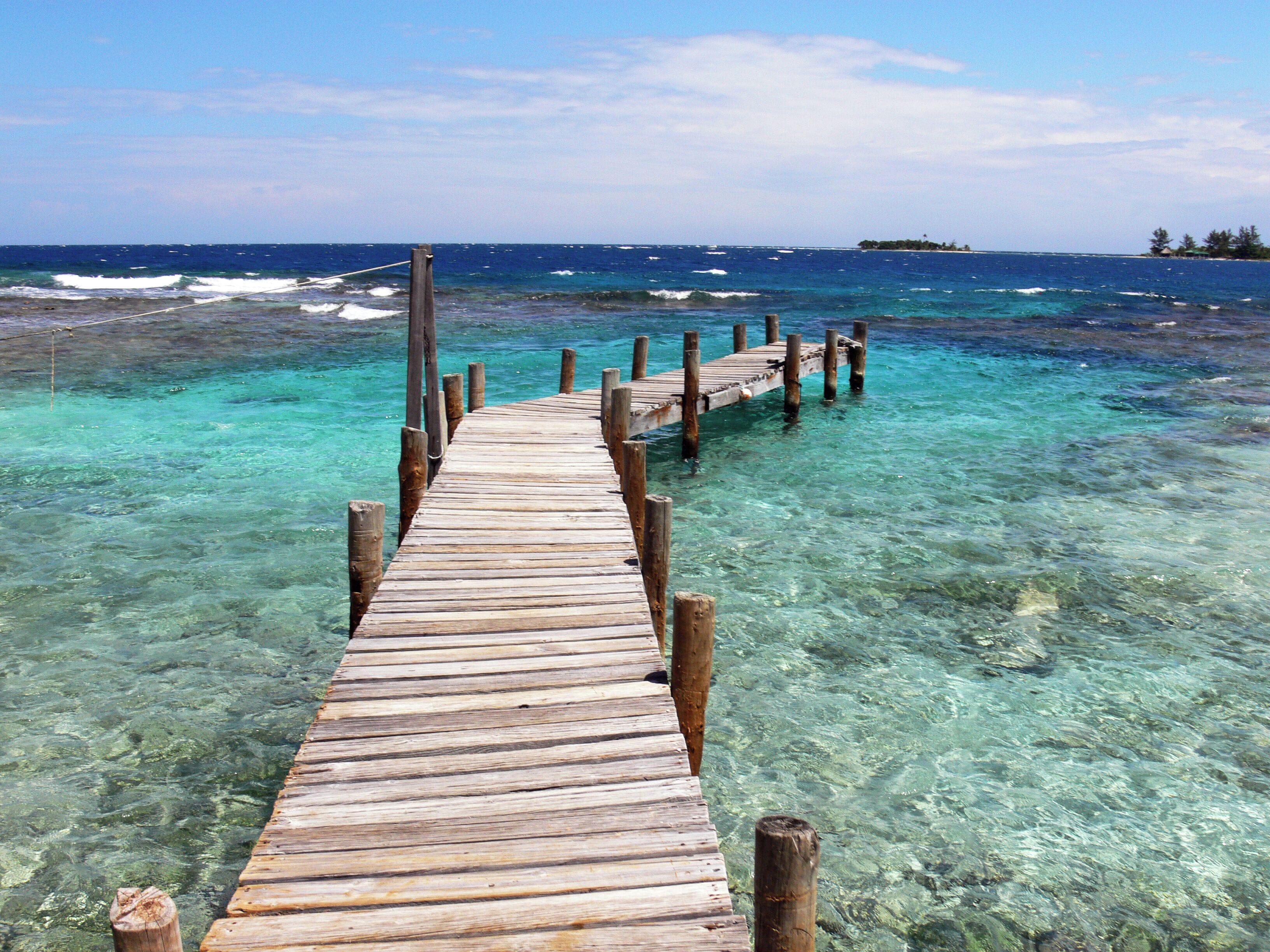 A wooden pier extending into clear turquoise waters with a distant island visible on the horizon.
