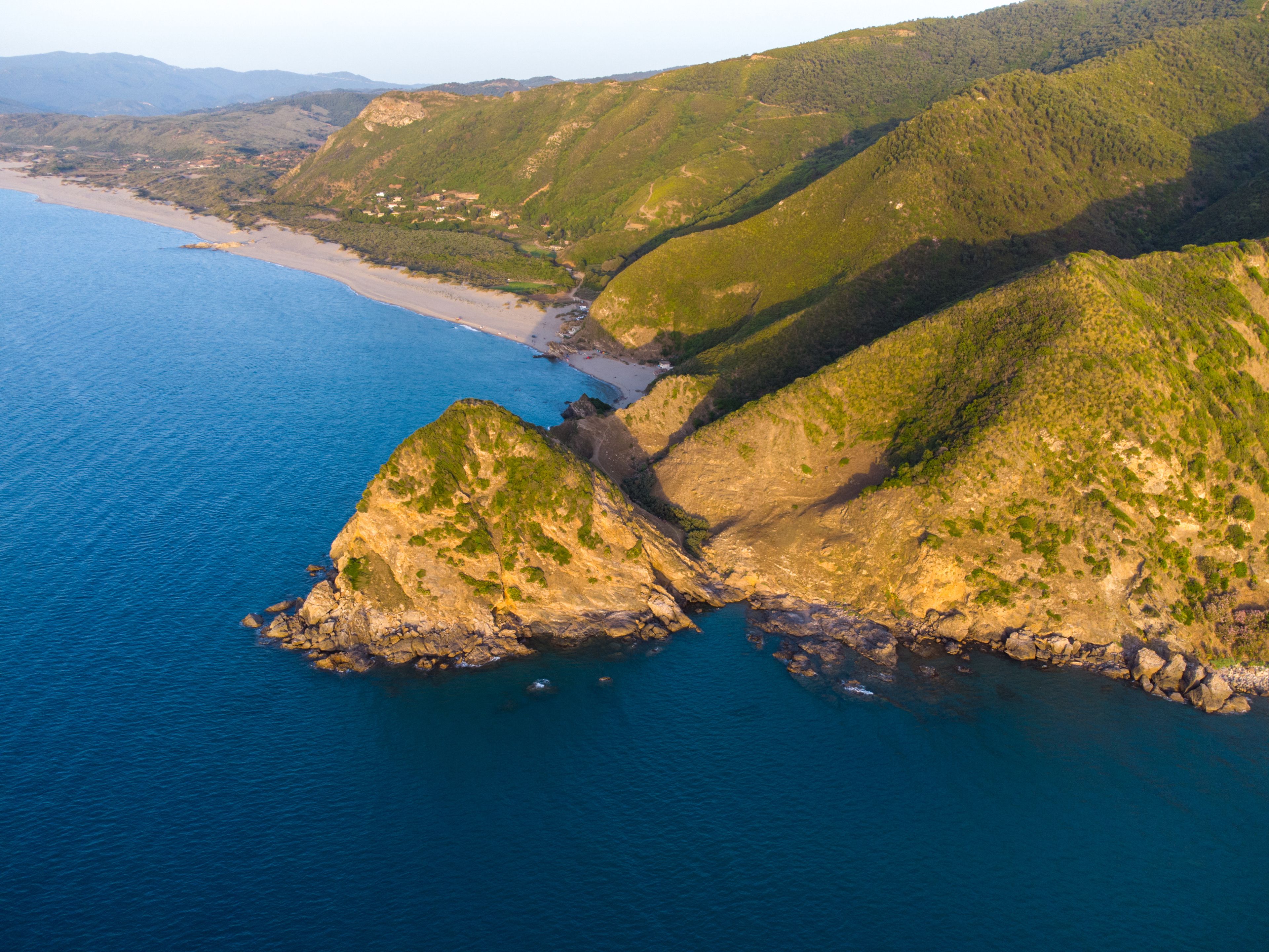 A coastal landscape with a rocky coastline, a sandy beach, and green hills.