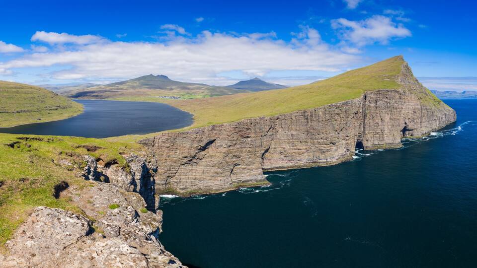 A coastal cliff with a steep drop into the sea, surrounded by grassy hills and distant mountains under a clear sky.