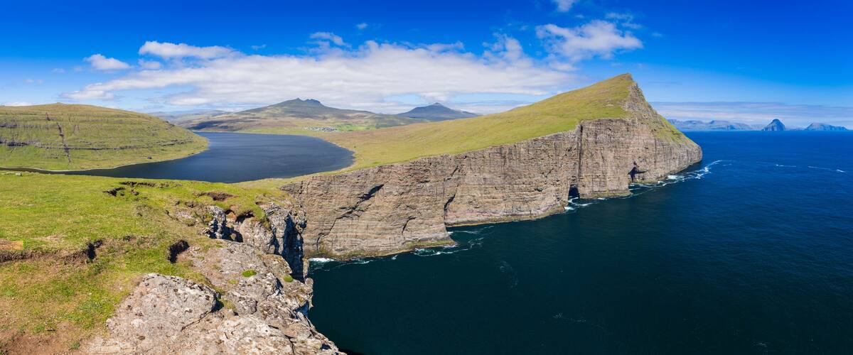 A coastal cliff with a steep drop into the sea, surrounded by grassy hills and distant mountains under a clear sky.