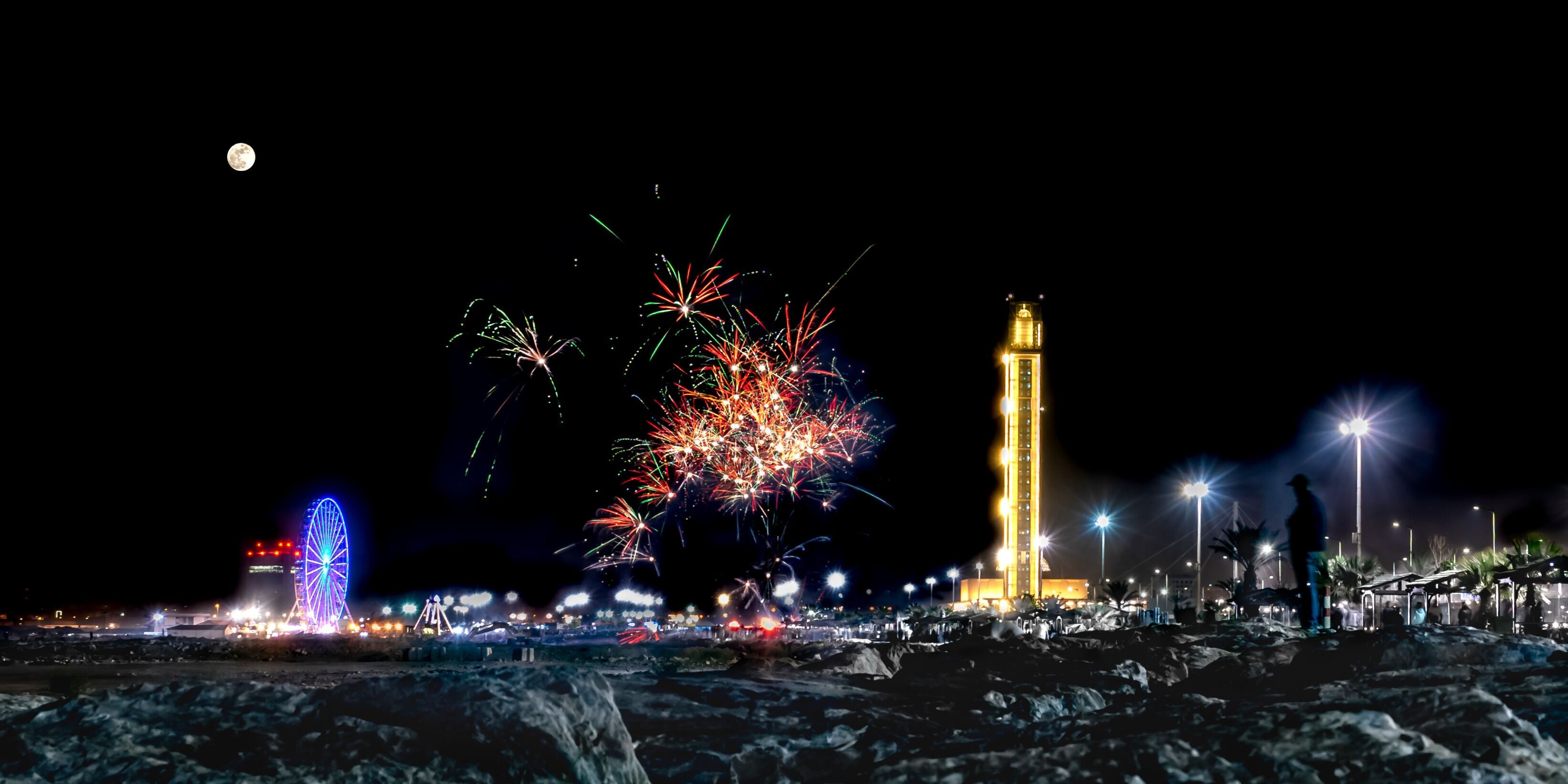 Algiers, Sablettes promenade fireworks. Djamaa el Djazaïr, full moon and colorful sparks at night. Ferris wheel, third-largest mosque and world's tallest minaret. A man stands facing the rocks. 