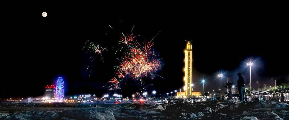 Algiers, Sablettes promenade fireworks. Djamaa el Djazaïr, full moon and colorful sparks at night. Ferris wheel, third-largest mosque and world's tallest minaret. A man stands facing the rocks.