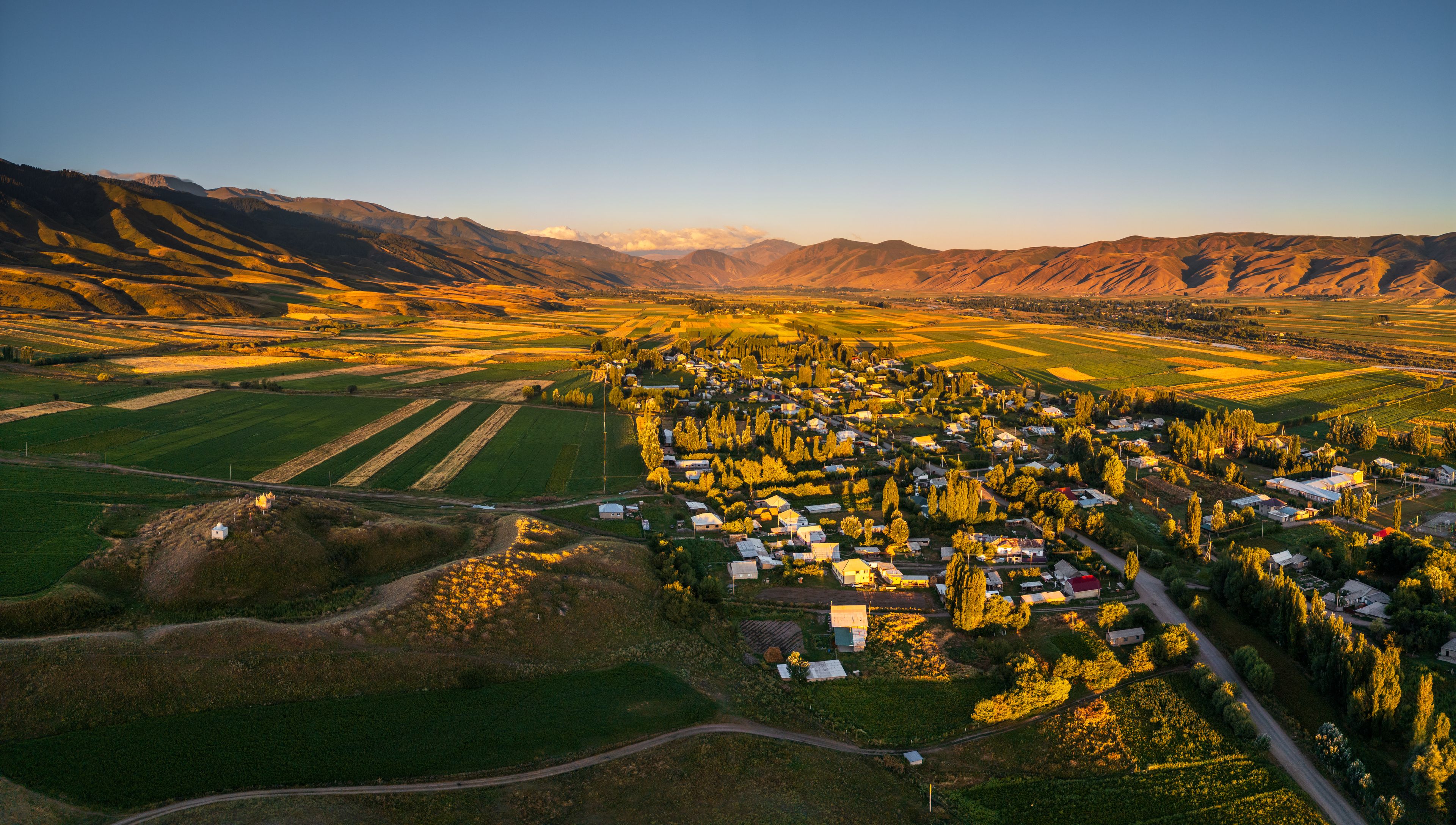 Aerial view of the Chon-Kemin valley village with agricultural fields and rolling mountains under the golden light of sunset in Kaindy, Chuy Province, Kyrgyzstan.