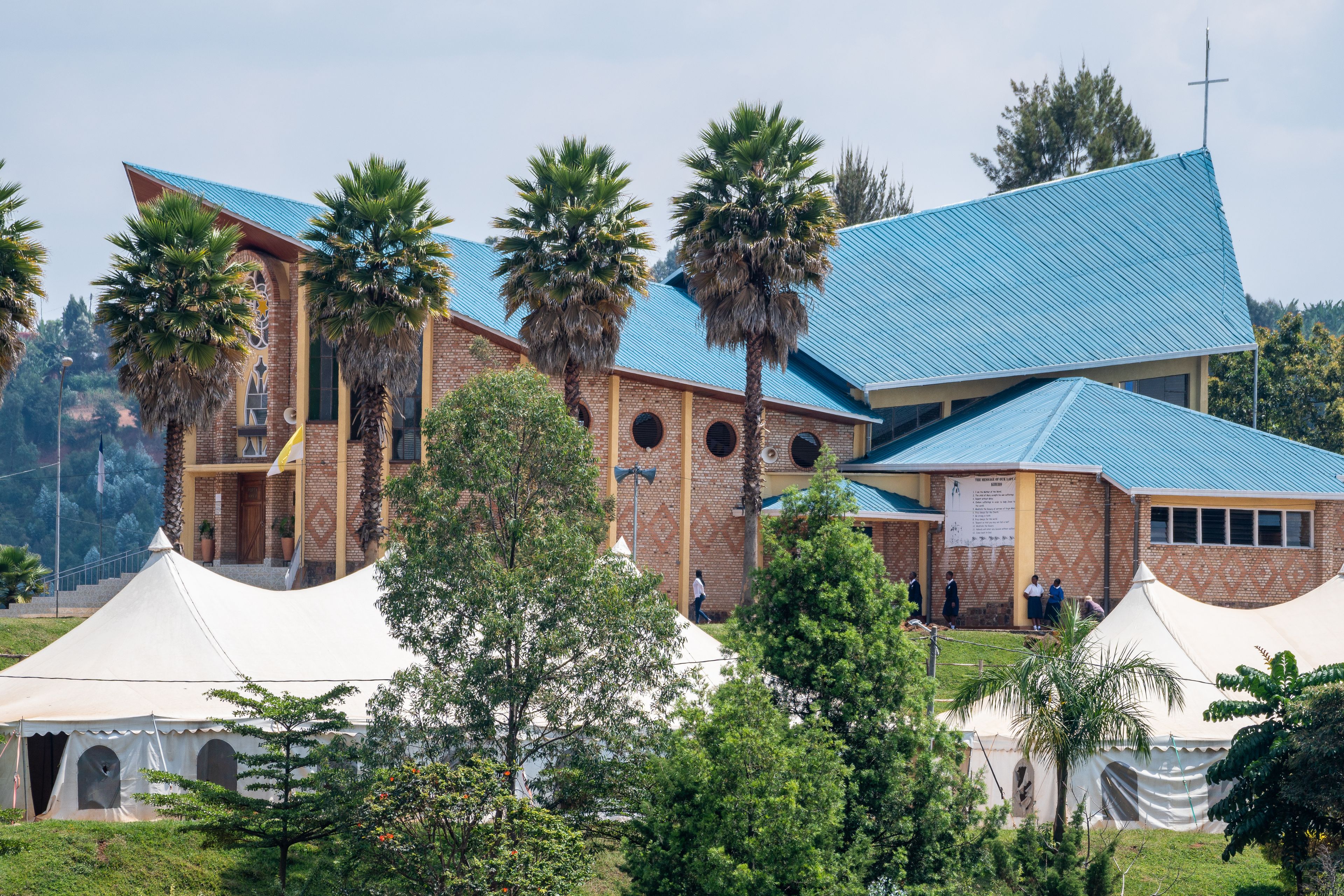 A church with a blue roof and brick walls, surrounded by palm trees and tents.