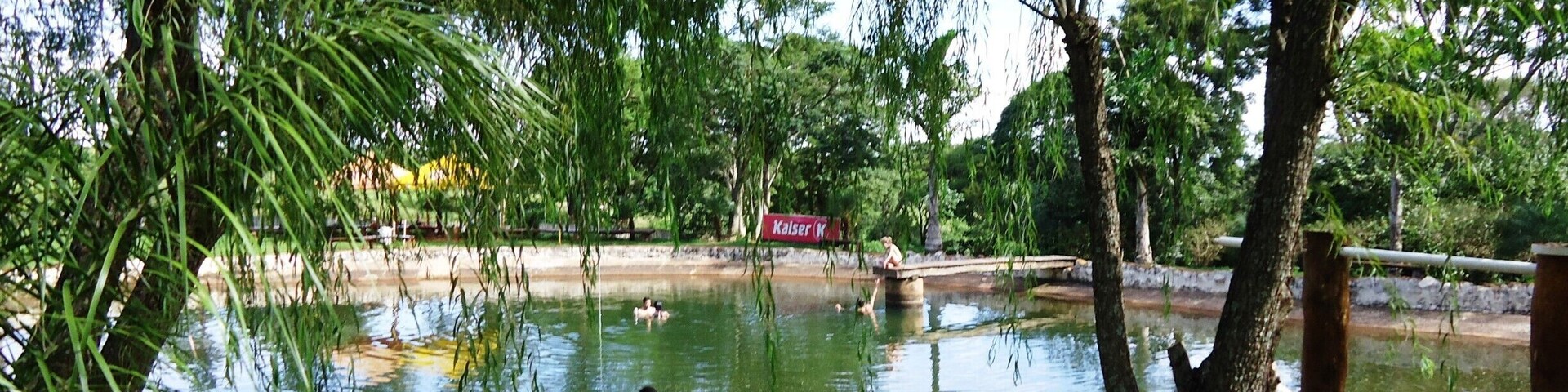 A pond with people swimming and walking around, surrounded by trees and a red sign in the background.