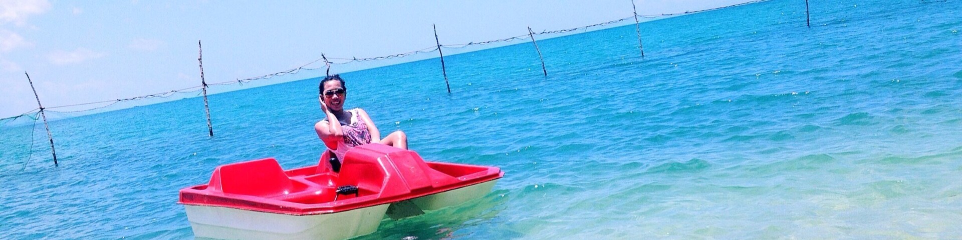 Uma pessoa em um barco vermelho e branco sobre águas azuis e claras, com uma cerca de arame farpado ao fundo.