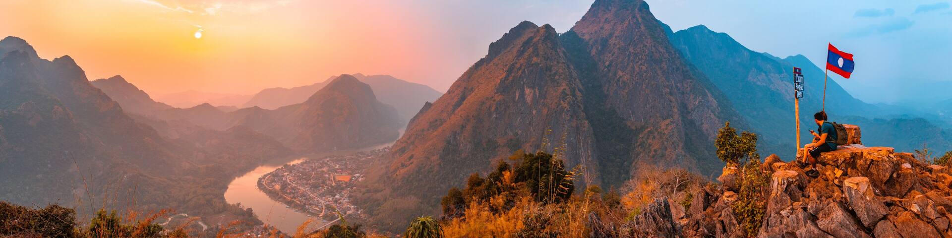Sunset panoramic view of couple of trekkers sitting on a rock on top of Nong Khiaw View Point with beautiful mountain and Nam Ou river in background, Luang Prabang Province, Northern Laos.