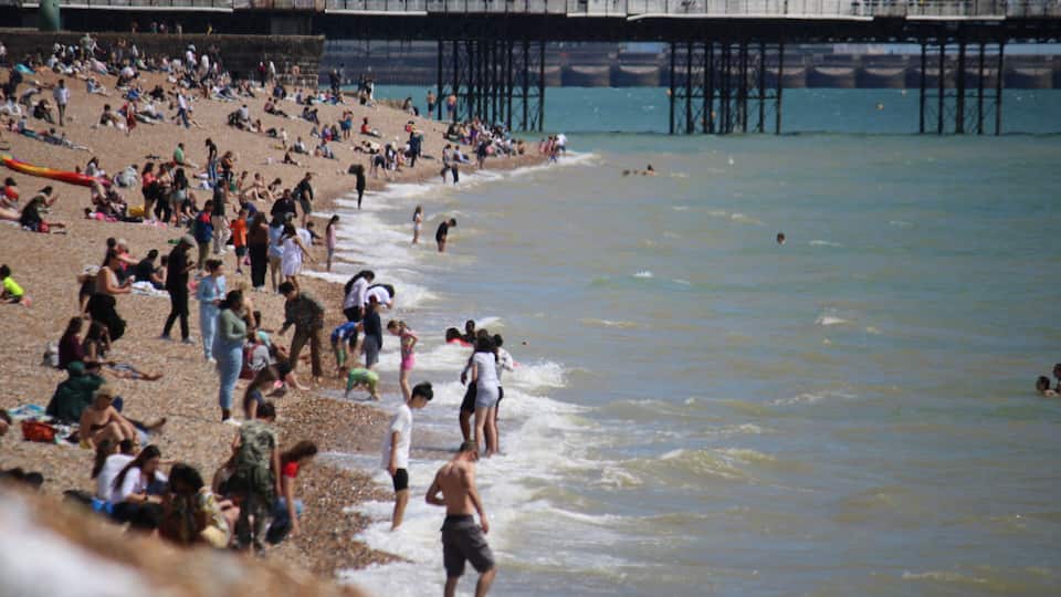 Brighton beach, East Sussex, UK - 08.08.2024: Brighton beach people sunbathing on shingle pebble beach sunny day with blue sky, Brighton pier behind