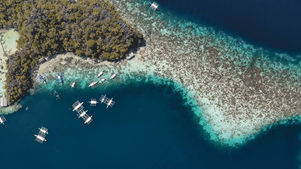 An aerial view of a tropical island with clear turquoise waters, a dense forested area, and several boats anchored near the shore.