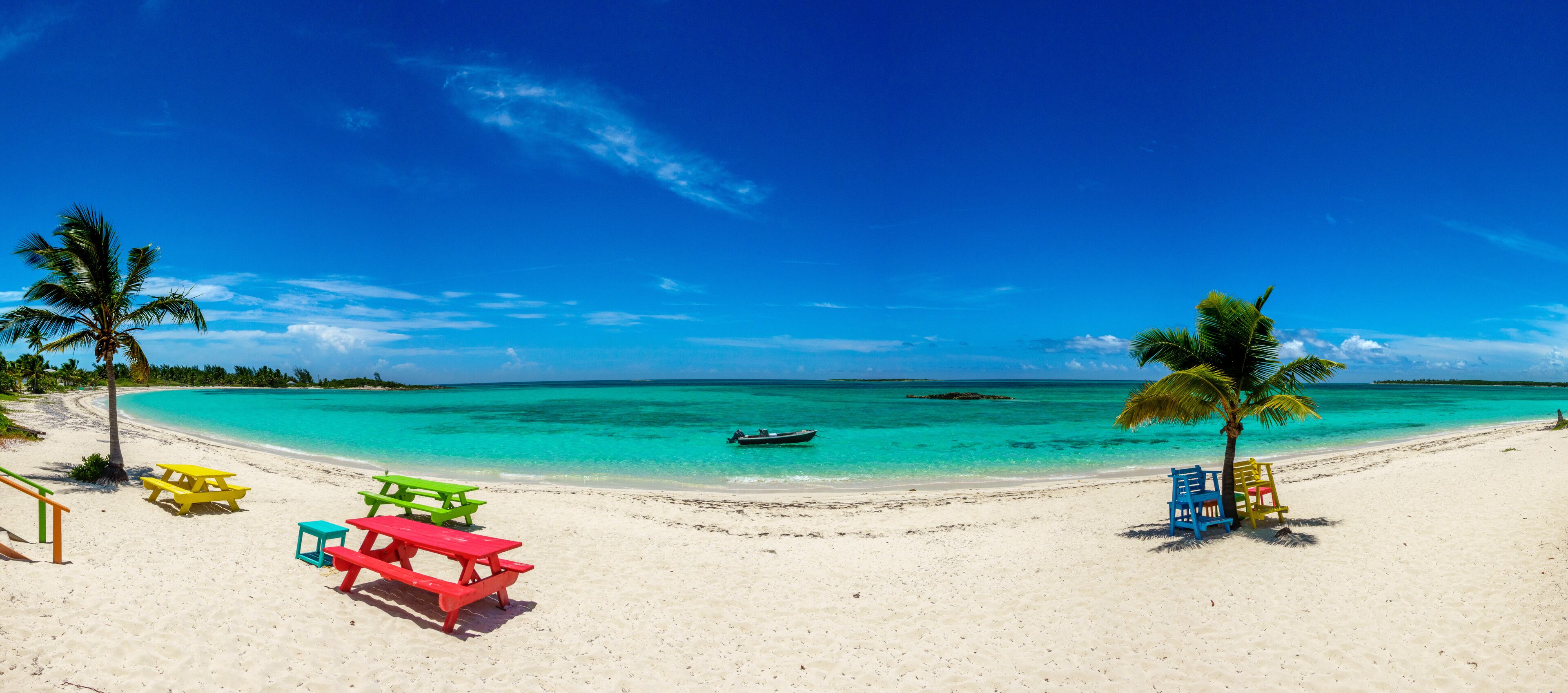 A beach with turquoise water, palm trees, and colorful picnic tables.