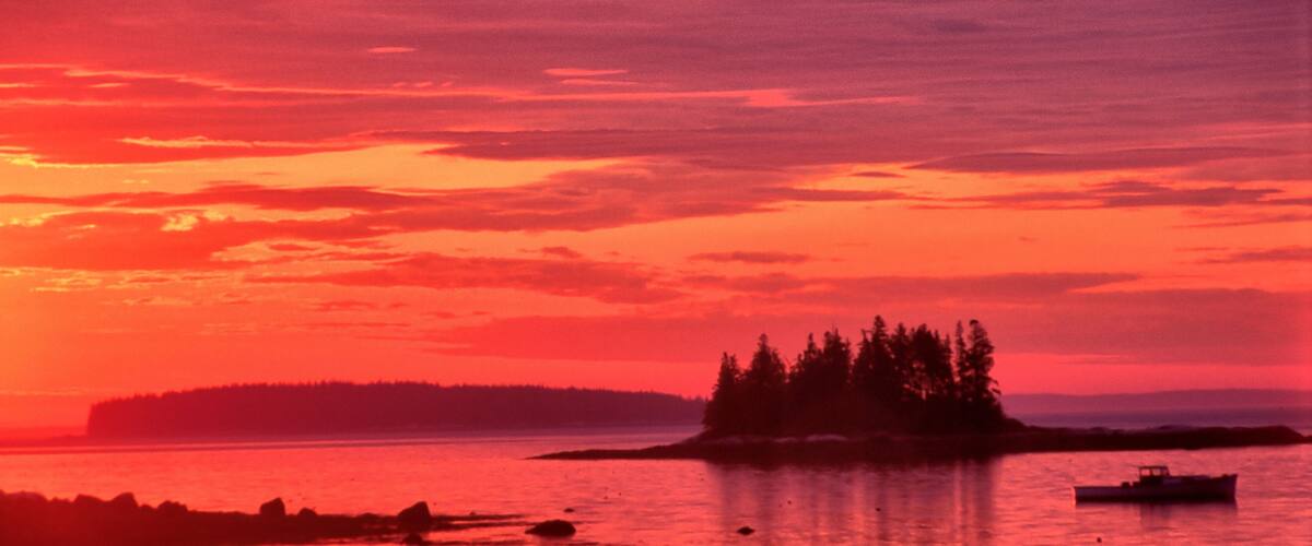 Vibrant red sky with sun about to rise over the horizon. Tranquil coastal scene near South Thomaston, Maine, with distant islands and lobster boat anchored in calm ocean.
