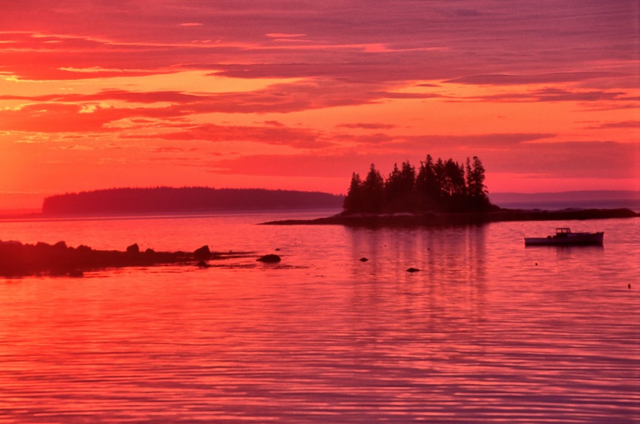 Vibrant red sky with sun about to rise over the horizon. Tranquil coastal scene near South Thomaston, Maine, with distant islands and lobster boat anchored in calm ocean.