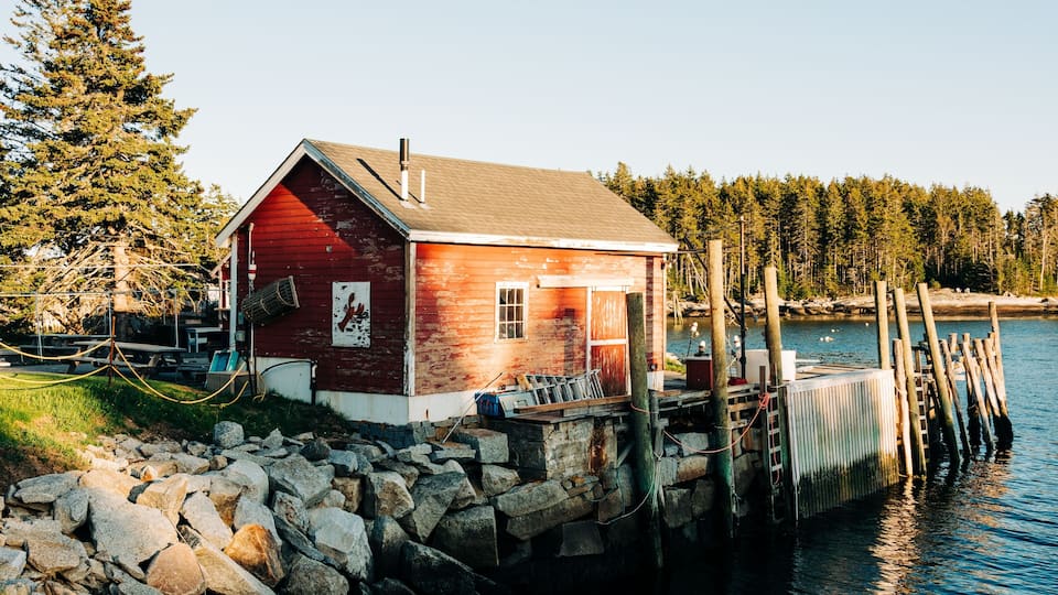 A wooden cabin by a rocky shore with a dock extending into calm water, surrounded by dense forest.