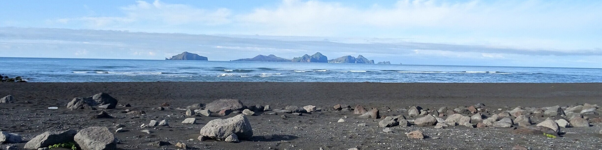 Uma praia rochosa com vegetação verde dispersa e ilhas ao longe.