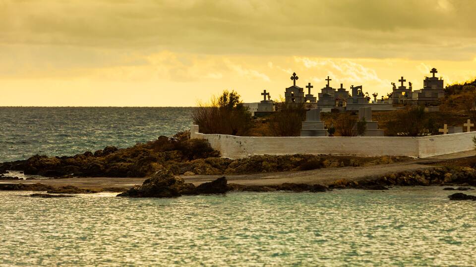 A coastal cemetery with numerous crosses and tombstones, surrounded by rocky terrain and the sea.