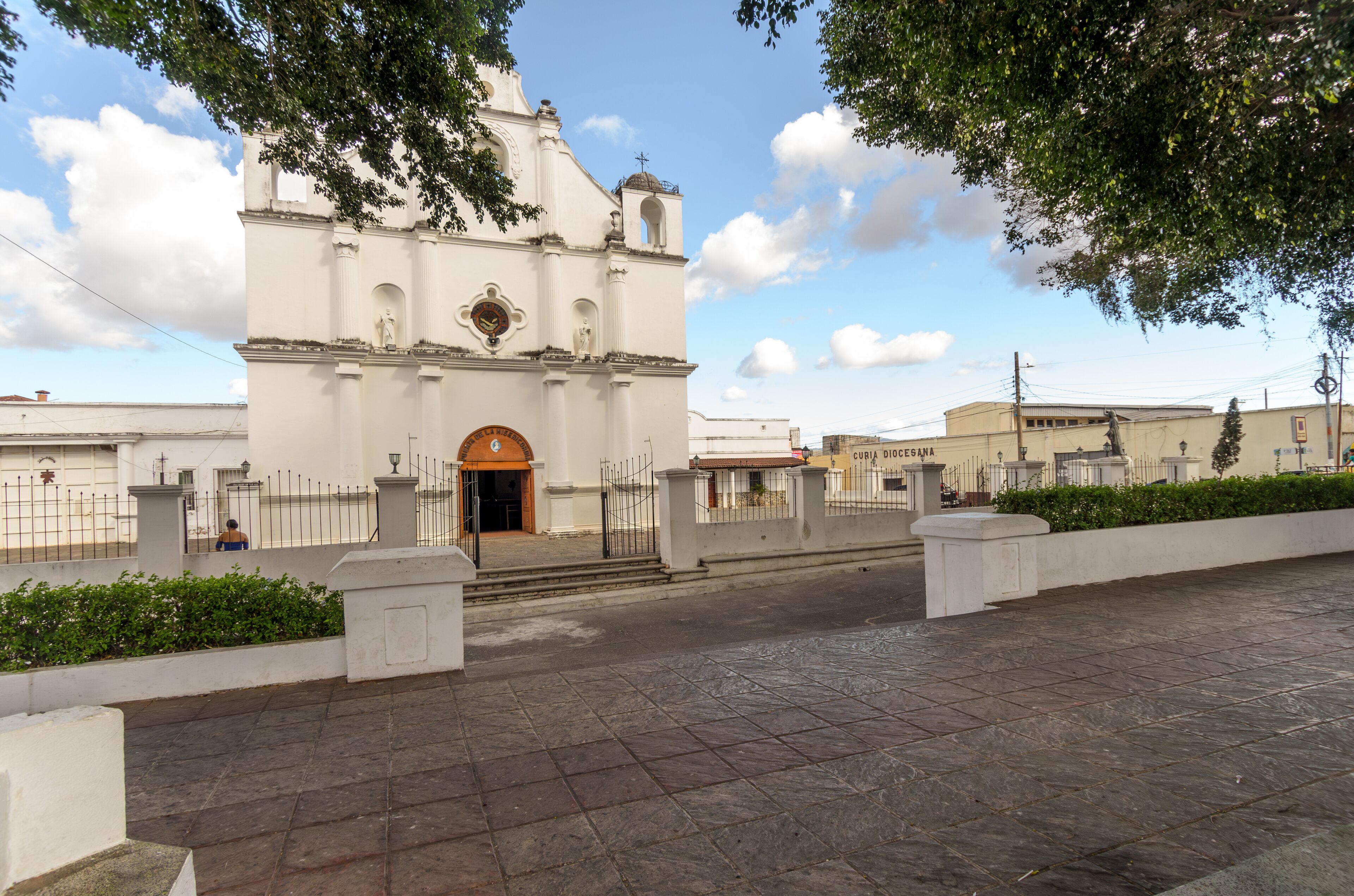 A white church with a clock tower and a cross on top, surrounded by a paved area and greenery.